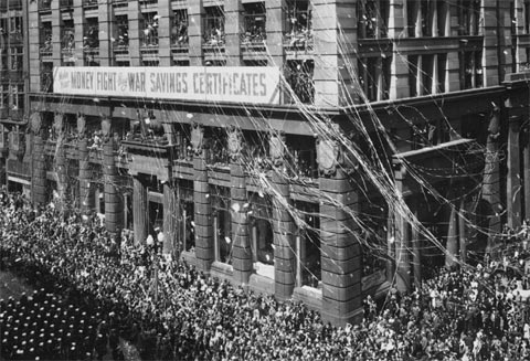 A rally for World War Two war savings certificates takes place outside the Commonwealth Bank. War savings certificates were designed by the Treasurer, Percy Spencer in early 1940 as a means to finance the war effort. Through issuing certificates the government was able to borrow money from the Australian public. From the outset, they proved very popular and a convenient form of investment of small sums of money. In a similar manner to the Diggers Loan of 1921, streamers and confetti were released onto the streets as part of the celebrations and campaign.