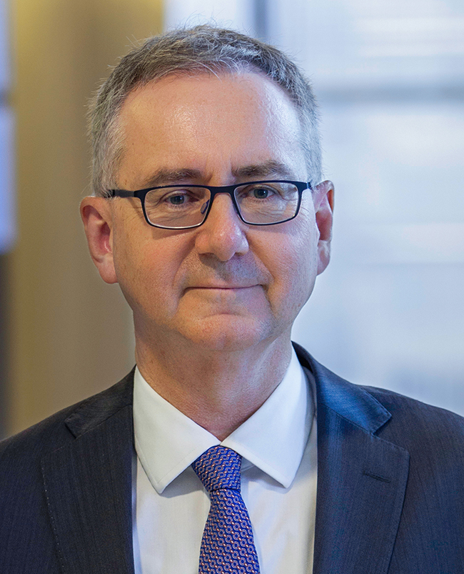 A headshot photograph of John, looking directly at the camera and smiling, wearing dark-framed glasses and a dark suit.