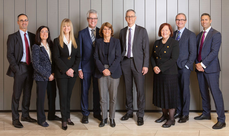 A photograph of the Payments System Board members standing in a row, dressed in suits looking directly at the camera and smiling. Michele Bullock is in the centre, wearing a dark jacket and grey pants.