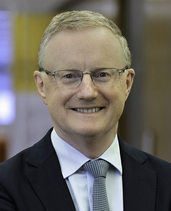 A headshot photograph of Philip, looking directly at the camera and smiling, wearing thin-framed glasses and a dark suit, white shirt and stripped tie.