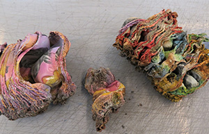 Three stacks of damaged banknotes that have been boiled. The first stack consists of $5 banknotes with damage around the edges and melted into a bowl shape. The second stack is a small lump of disfigured and boiled $5 banknotes. The third stack contains a mix of warped $5, $10, $20, $50 and $100 banknotes stuck together with significant damage.